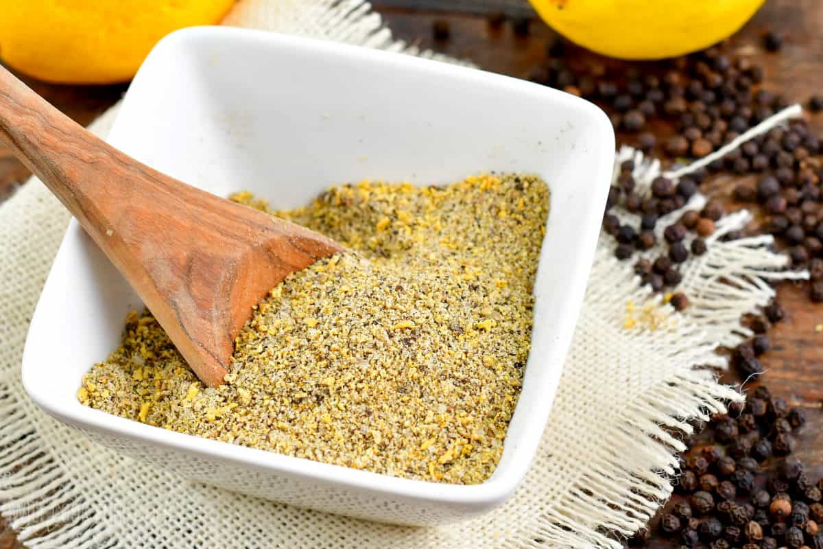 Lemon pepper seasoning in a bowl with a wood spoon on a table cloth with peppercorns around it.