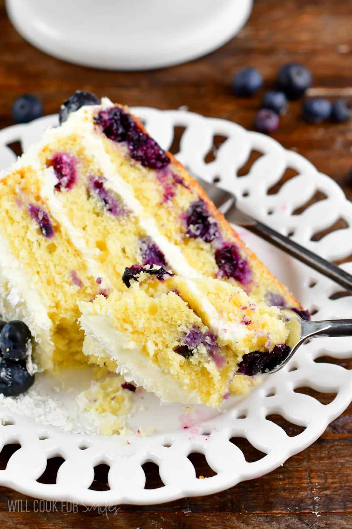 Using a fork to scoop up some of a slice of blueberry cake that is on a plate.