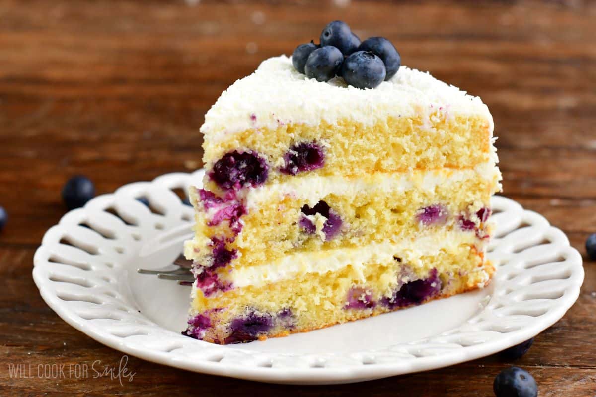 A slice of blueberry cake on a plate with blueberries on top table around it.