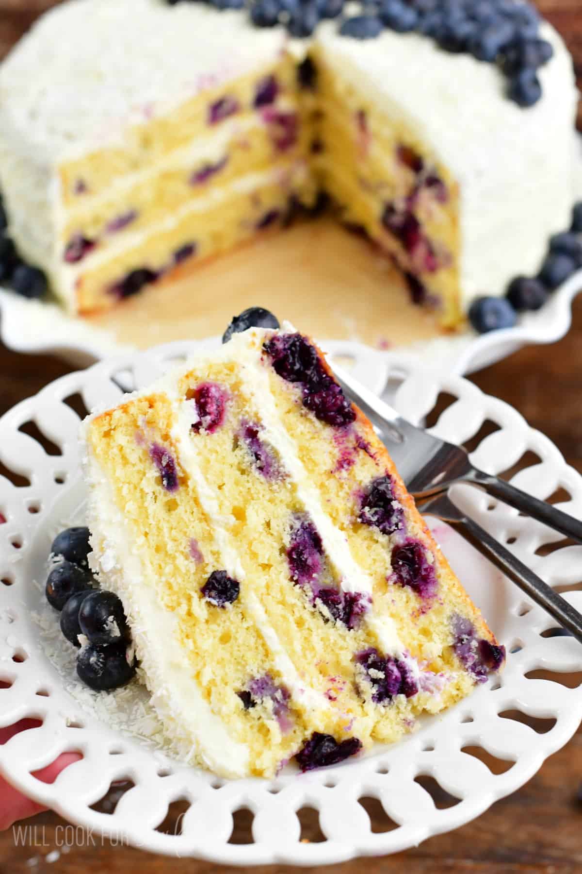 A slice of blueberry cake on a plate with the rest of the cake next to it on a cake stand.