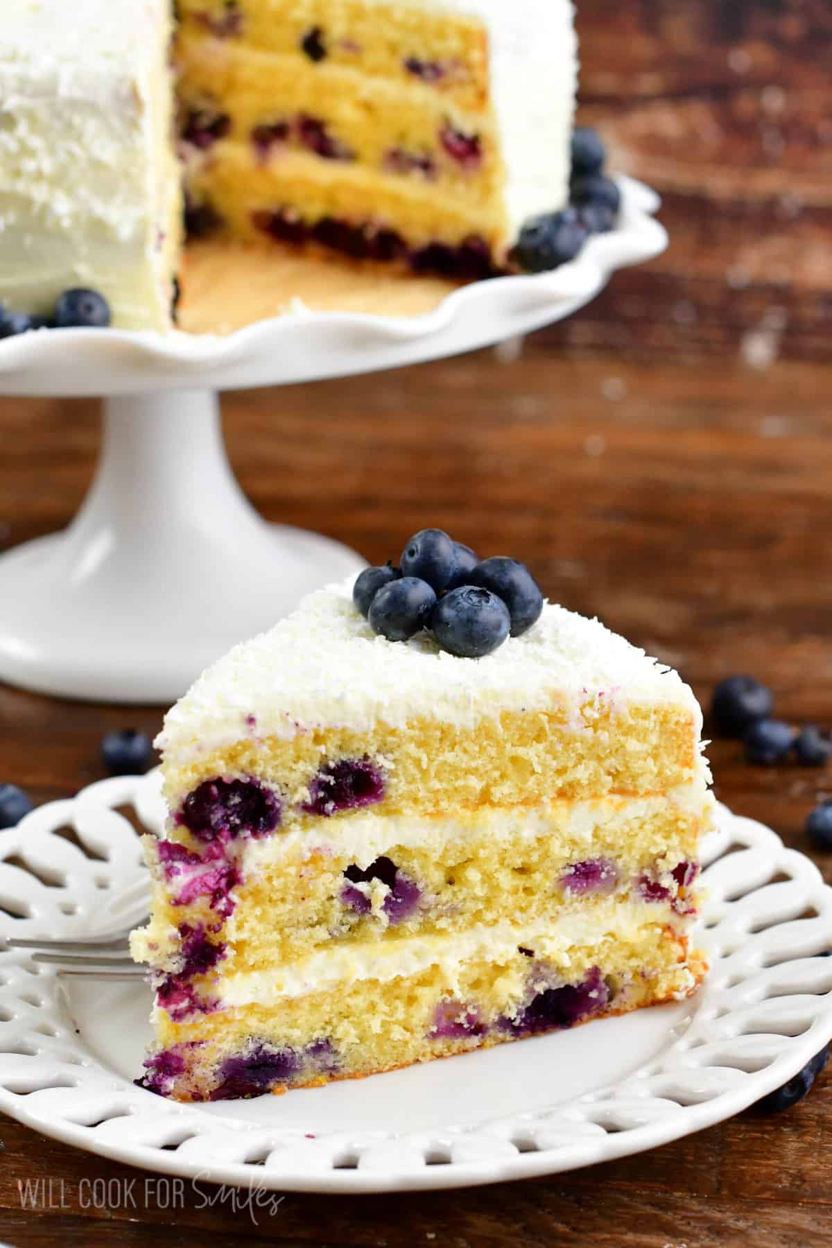 A slice of blueberry cake on a plate next to the cake stans with the rest of the cake.