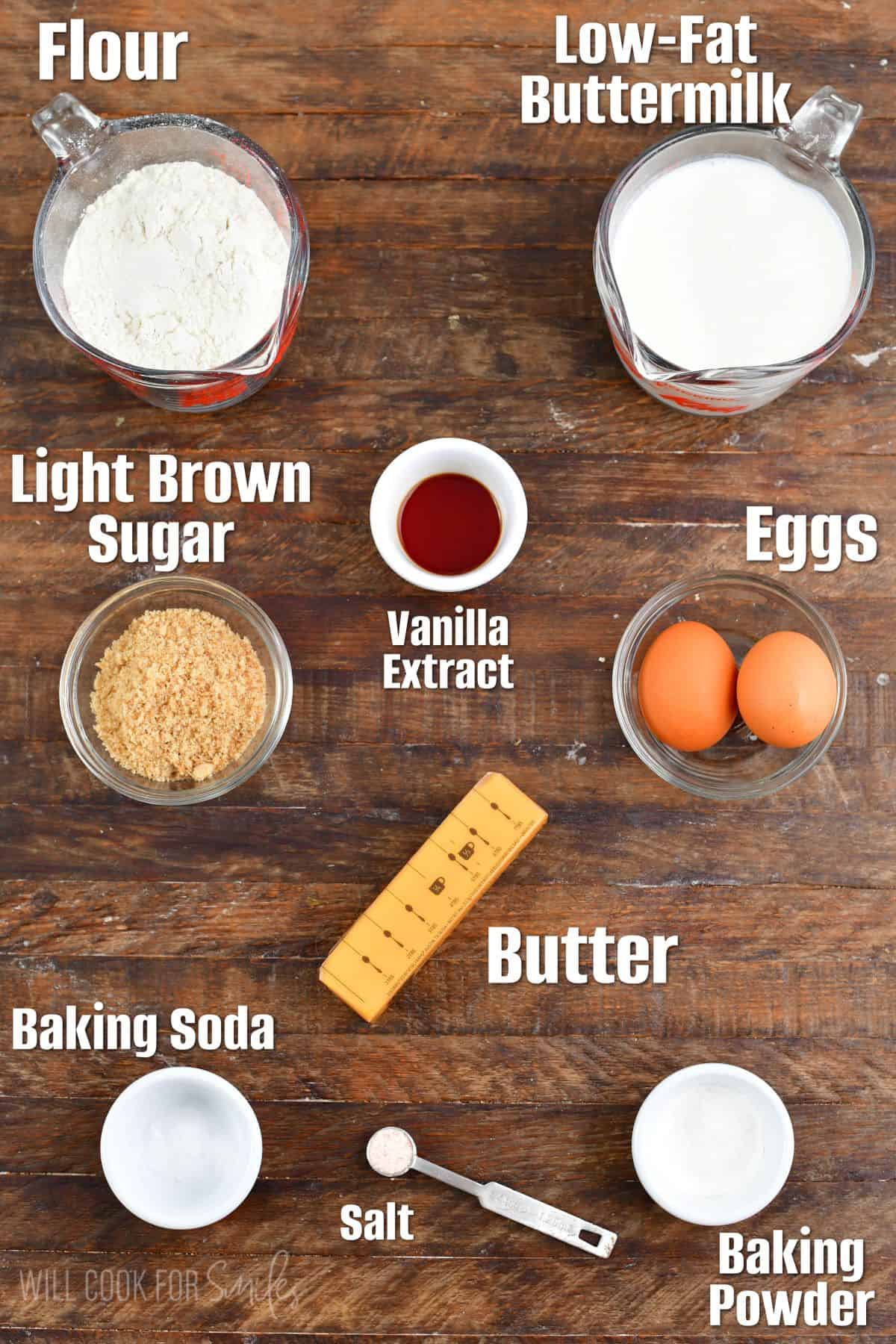 labeled ingredients to make waffles in measuring cups on a wooden surface.