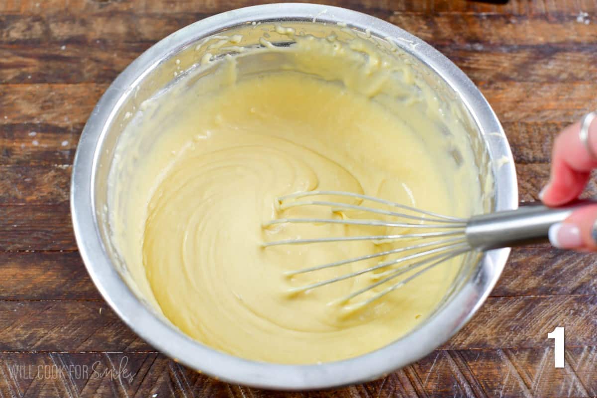whisking a light colored waffle batter in a metal mixing bowl.