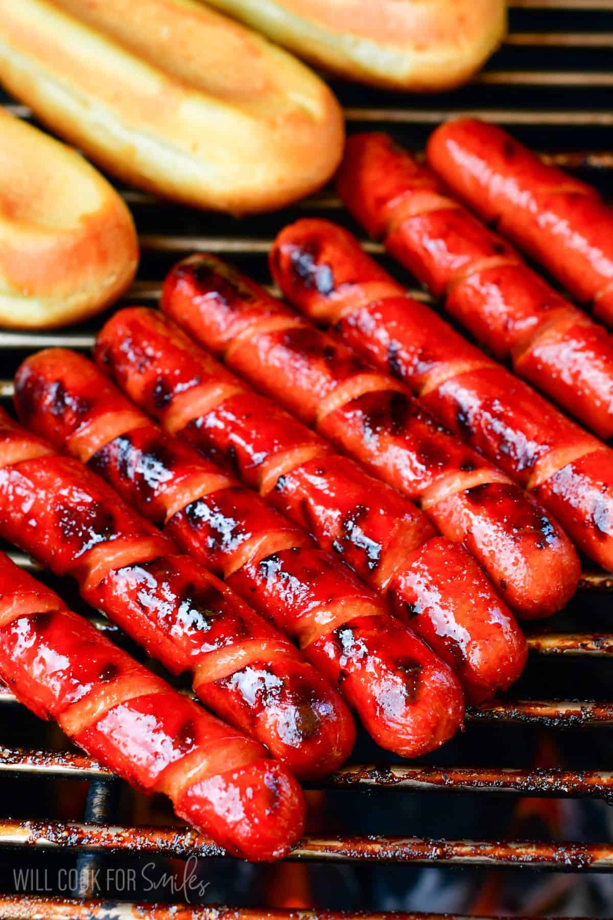 Sriracha Honey Hot Dogs cooking on the grill with the hotdog buns next to them.