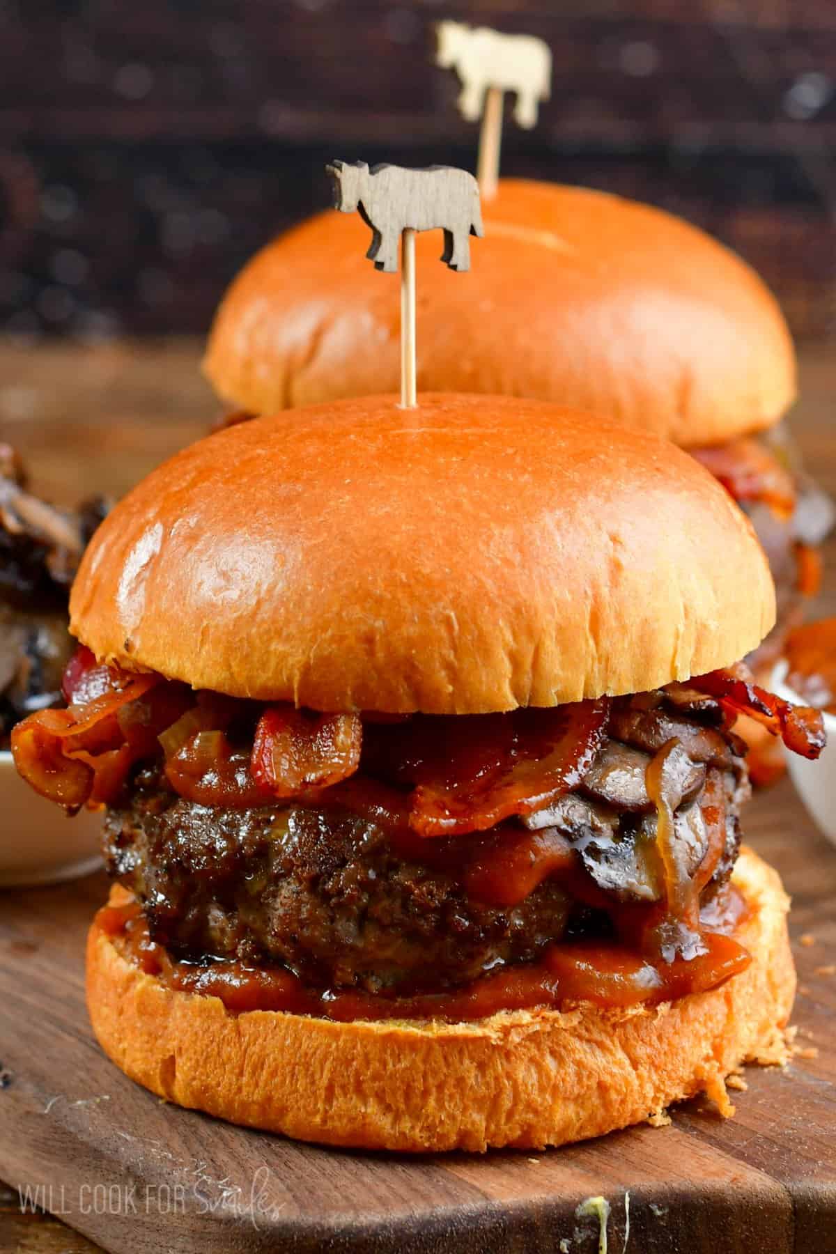 Two burgers with sauteed mushrooms, bacon, and whiskey sauce on a bun on a wood surface.