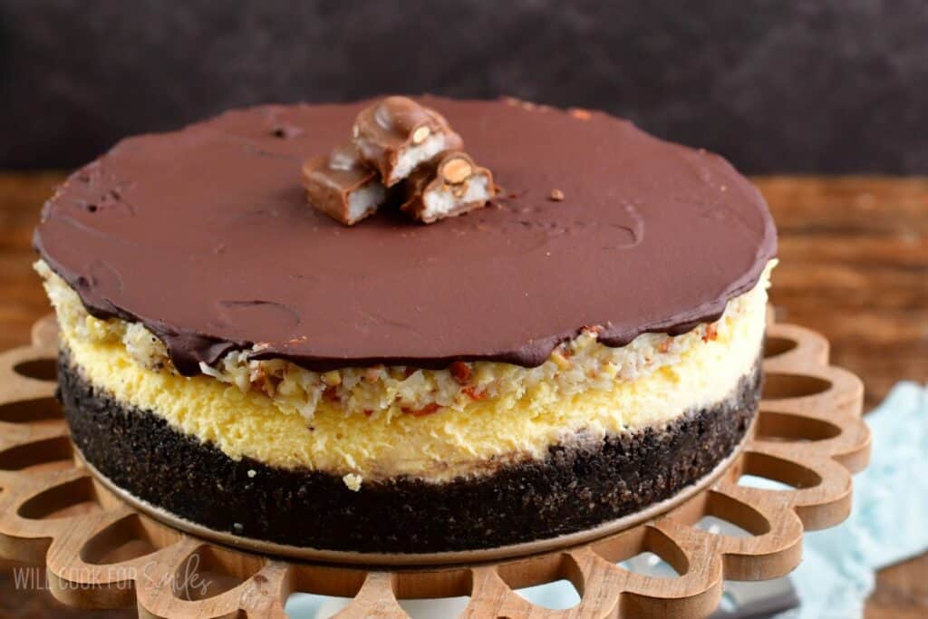 Whole almond joy cheesecake on a wood cake stand with a cut up almond joy on top.