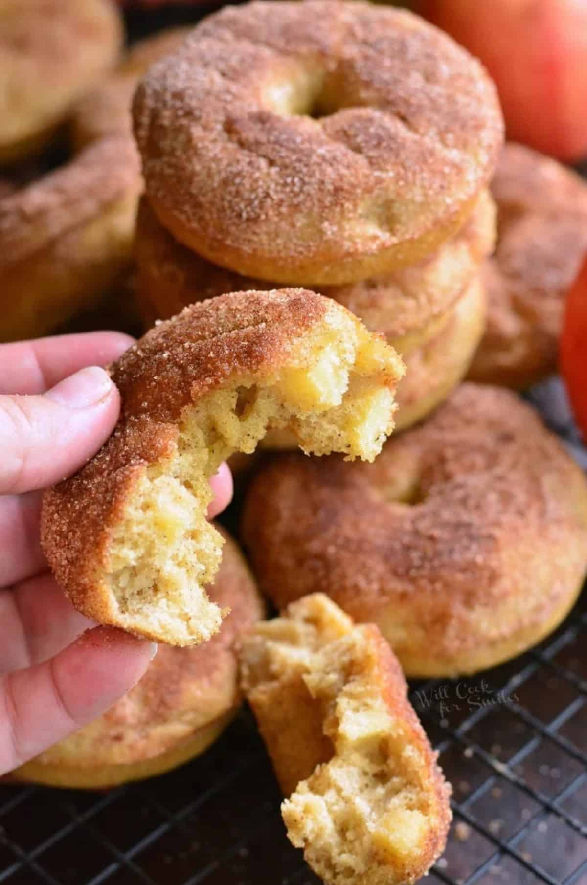 Apple baked donuts stacked up on a wire rack and holding one that has been torn in half.