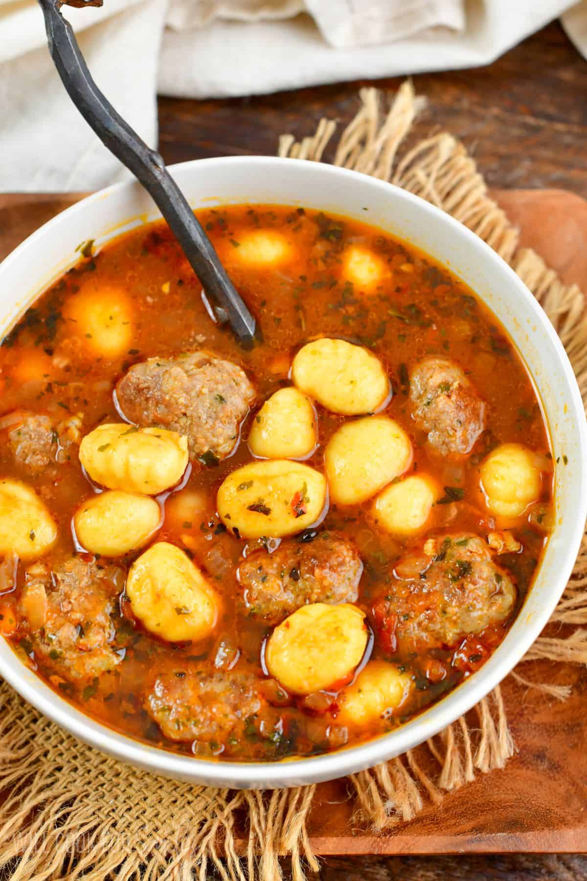 Italian meatballs and gnocchi soup in a bowl with a spoon on wooden plate.