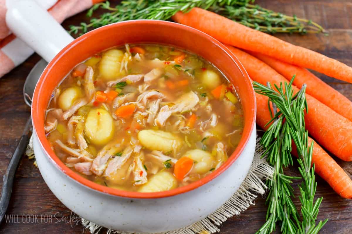 a bowl filled with clear chicken gnocchi soup with veggies and rosemary around.