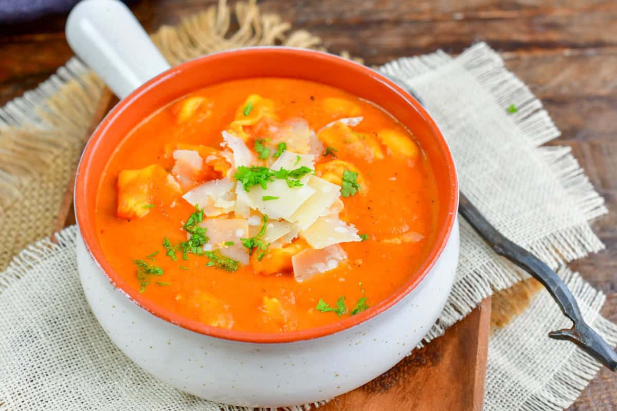 bowl of roasted red pepper soup on a wooden plate with a spoon next to it.