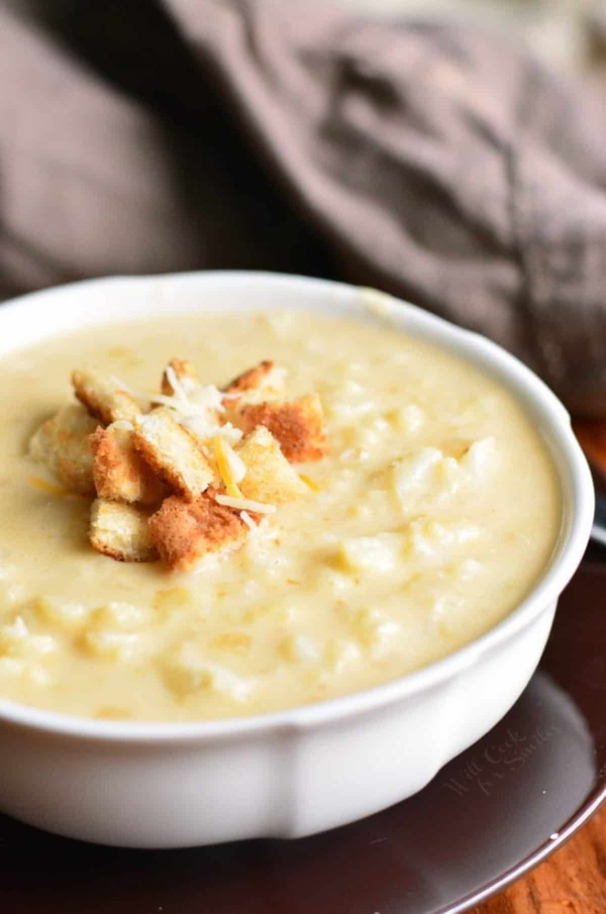 white bowl full of cheesy cauliflower soup topped with small bread croutons and shredded cheese.