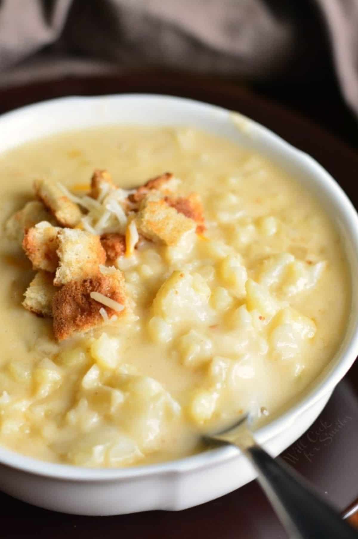 bowl full of cauliflower cheese soup topped with croutons and cheese with a spoon in it.