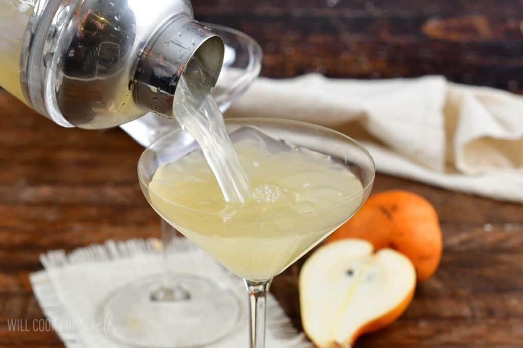 pouring a light yellow pear martini into a martini glass.