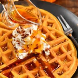 Pouring syrup over two waffles on a plate with whipped cream and nuts on top.