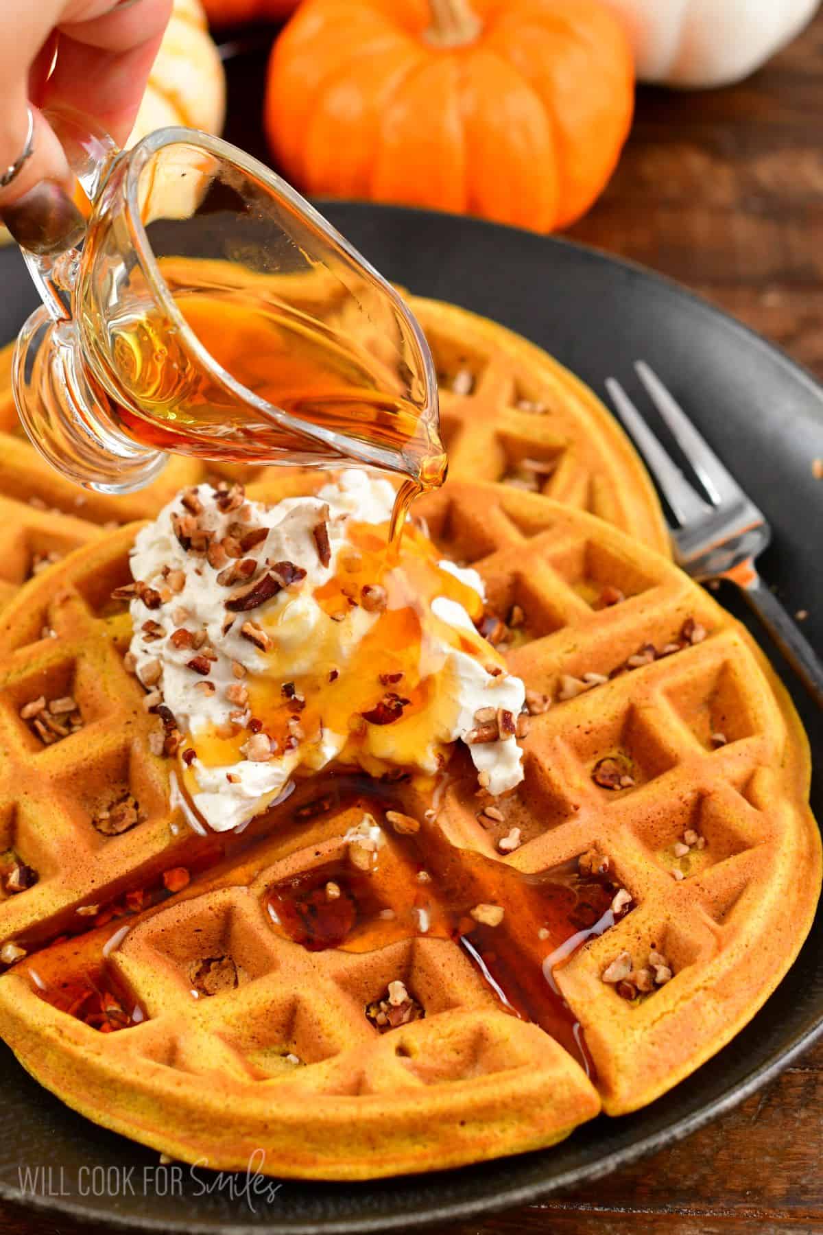 Pouring syrup over two waffles on a plate with whipped cream and nuts on top.