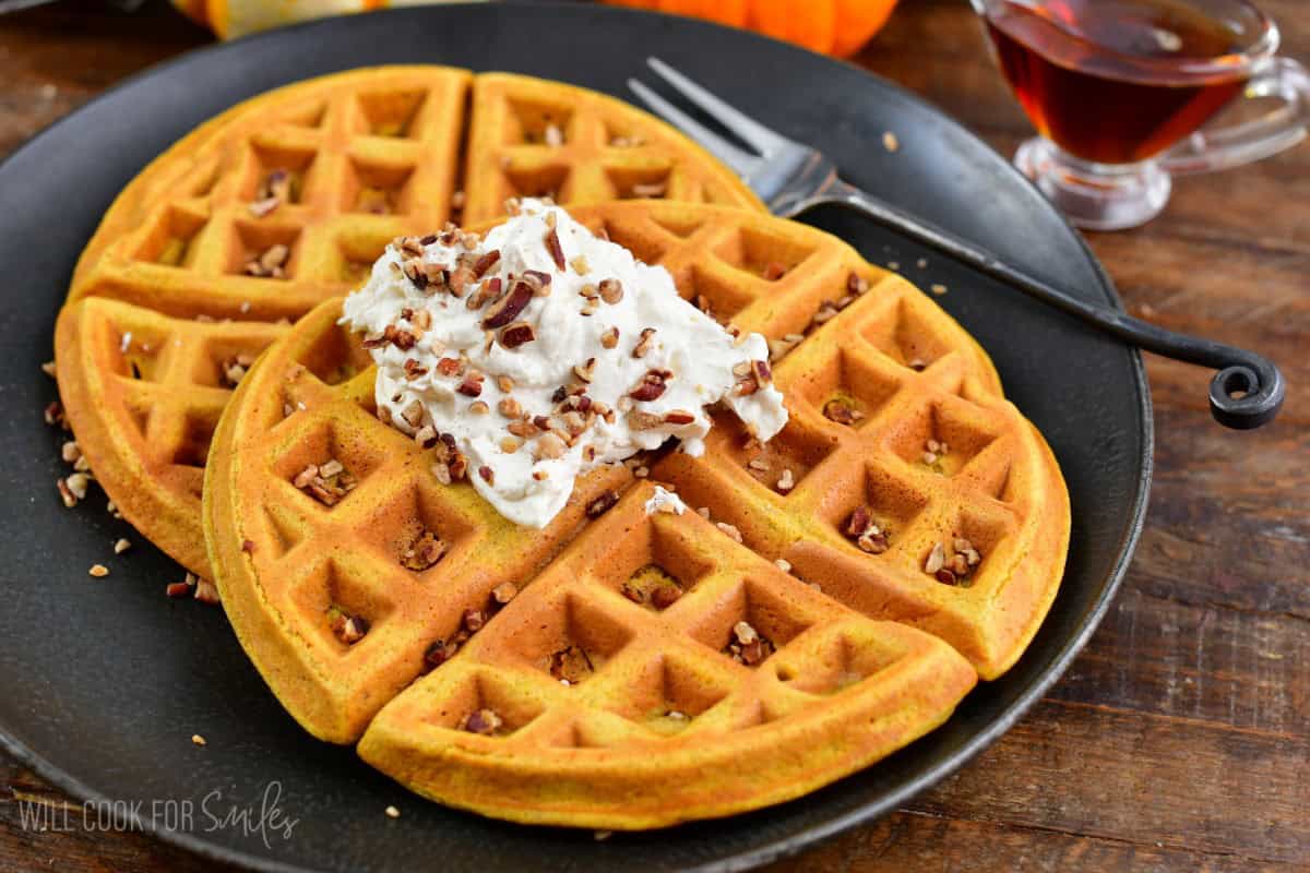 Two pumpkin waffles stacked on plate with whipped cream and nuts on top.