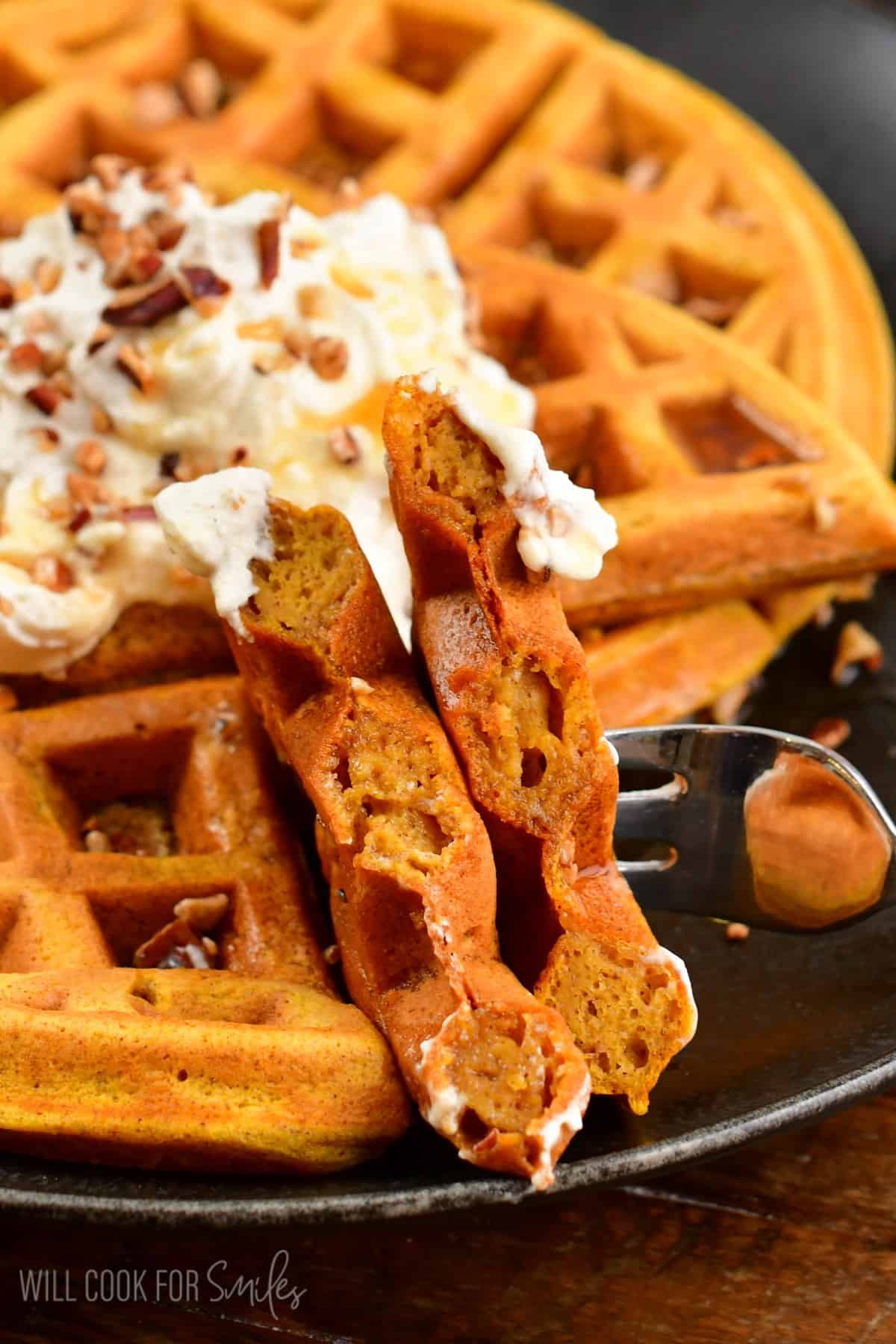 Pumpkin waffles with whipped cream and nuts on top and a bite on a fork.