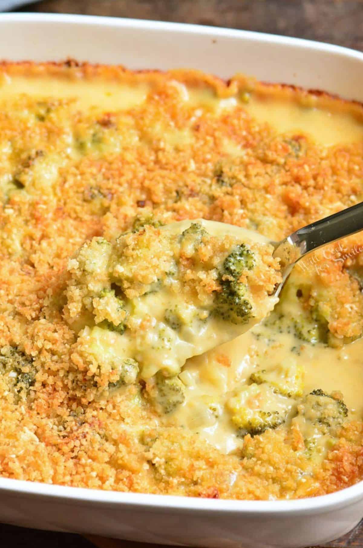 spooning some broccoli cheese casserole out of the baking dish with silver spoon.