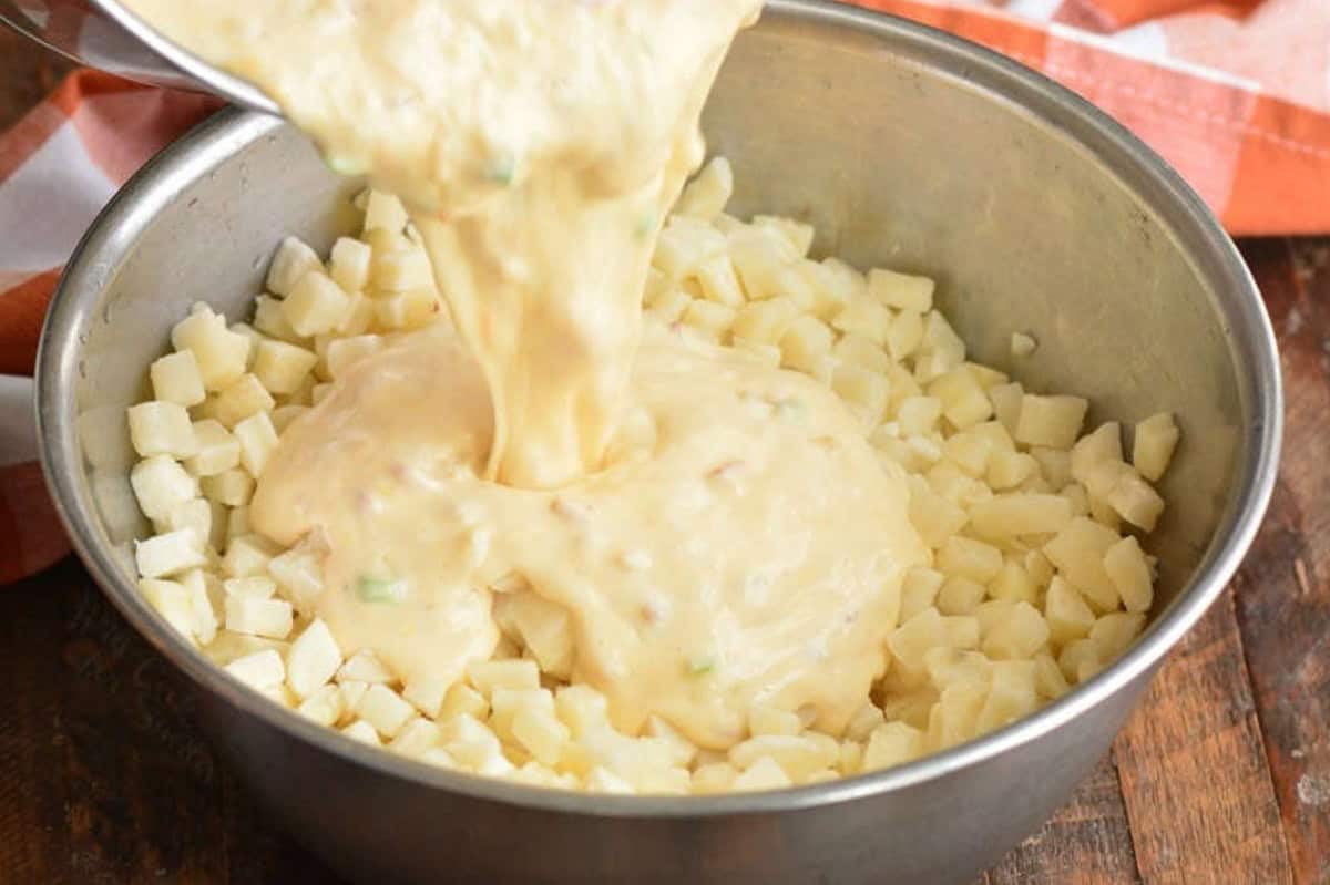 Adding cheese sauce to diced potatoes in a metal bowl.