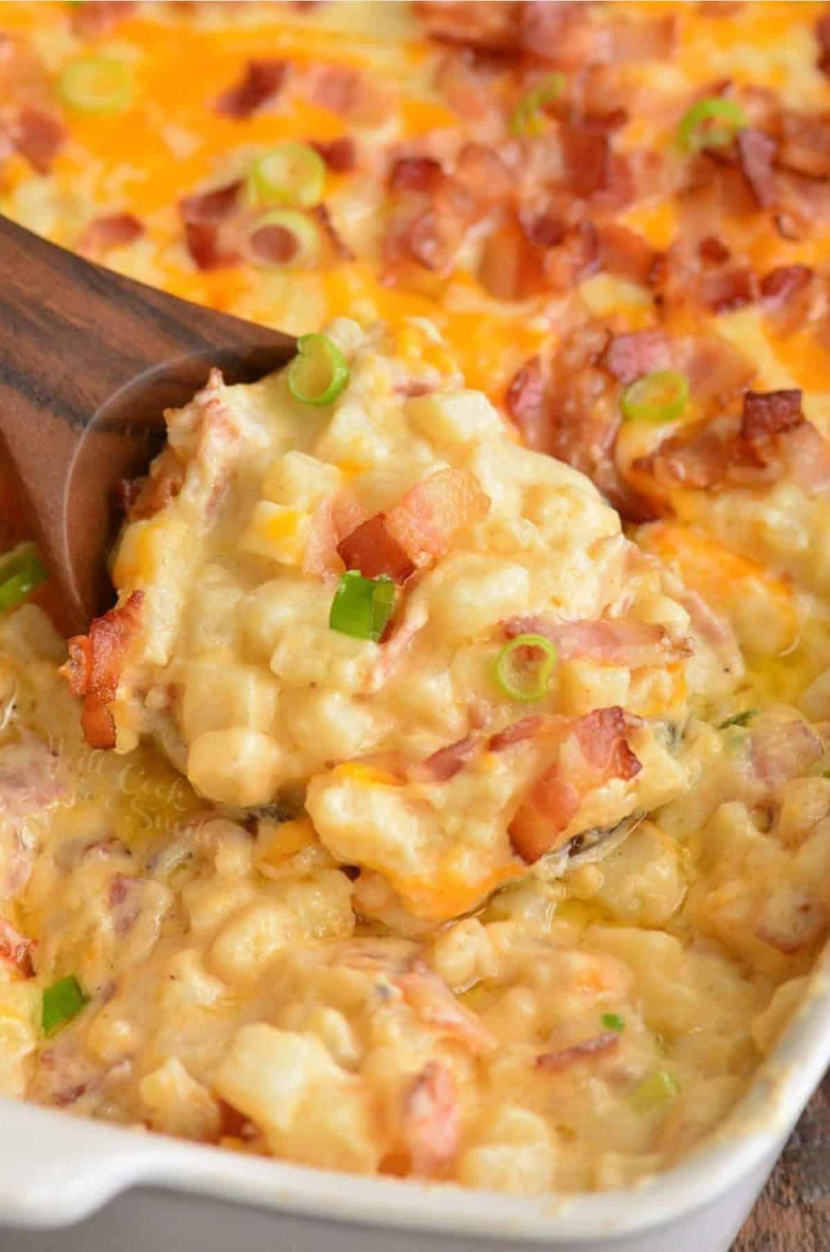 Baked loaded cheesy potato casserole in a baking dish with a wood spoon scooping some out.