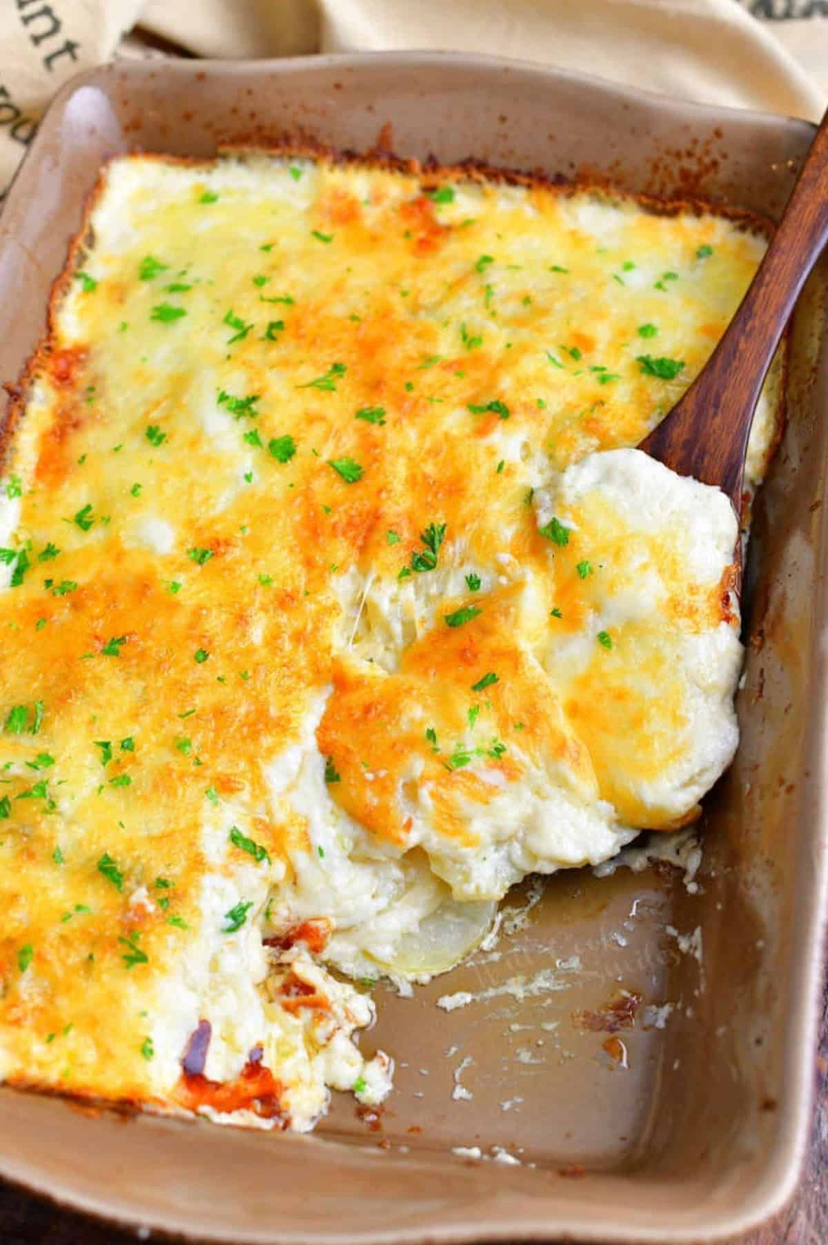 scalloped poatoes in a baking dish with a some taken out and a wooden spoon scooping some.