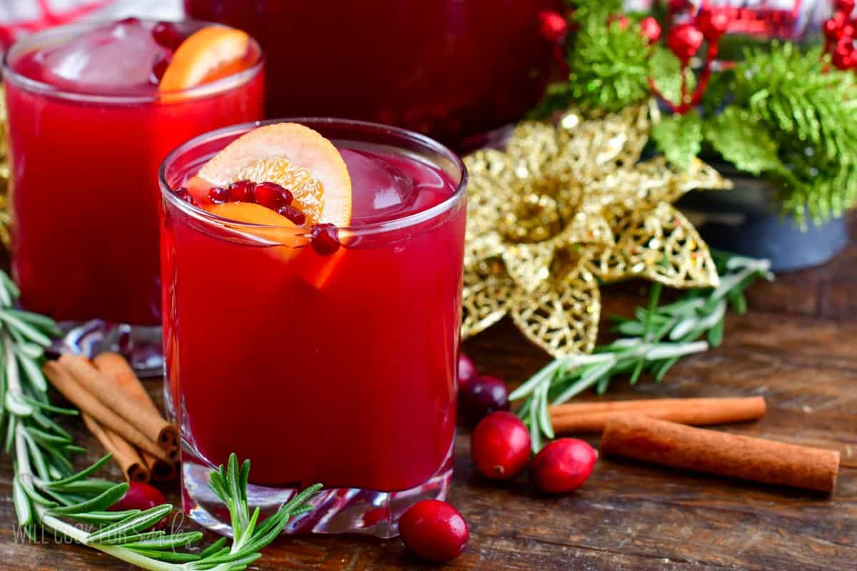 Christmas punch in a glass with another behind and rosemary and cranberries and cinnamon sticks around.