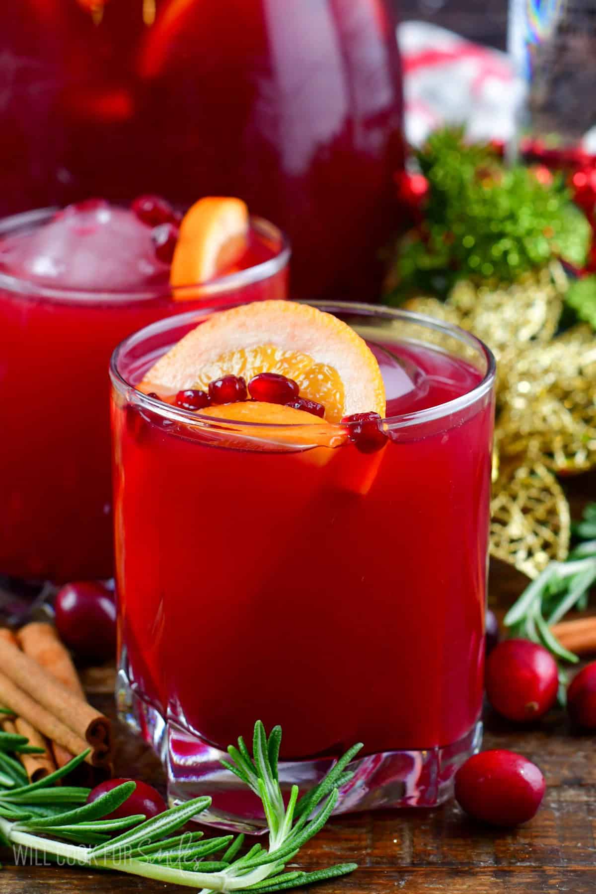 Christmas punch in a glass with another behind with rosemary, cranberries and cinnamon sticks around.