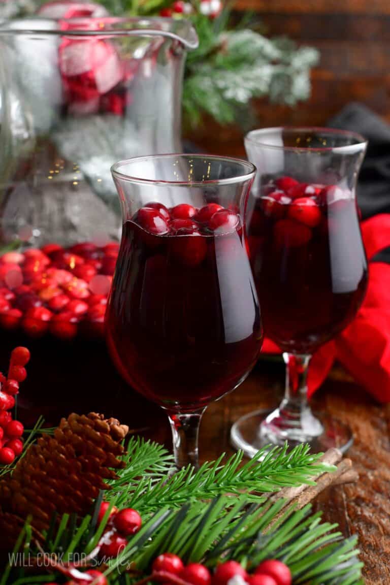 two glasses of cranberry sangria with cranberries and a pitcher behind.