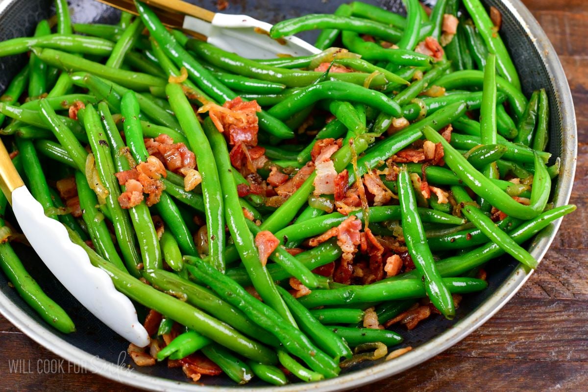 sauteed green beans with bacon in a cooking pan with tongs.