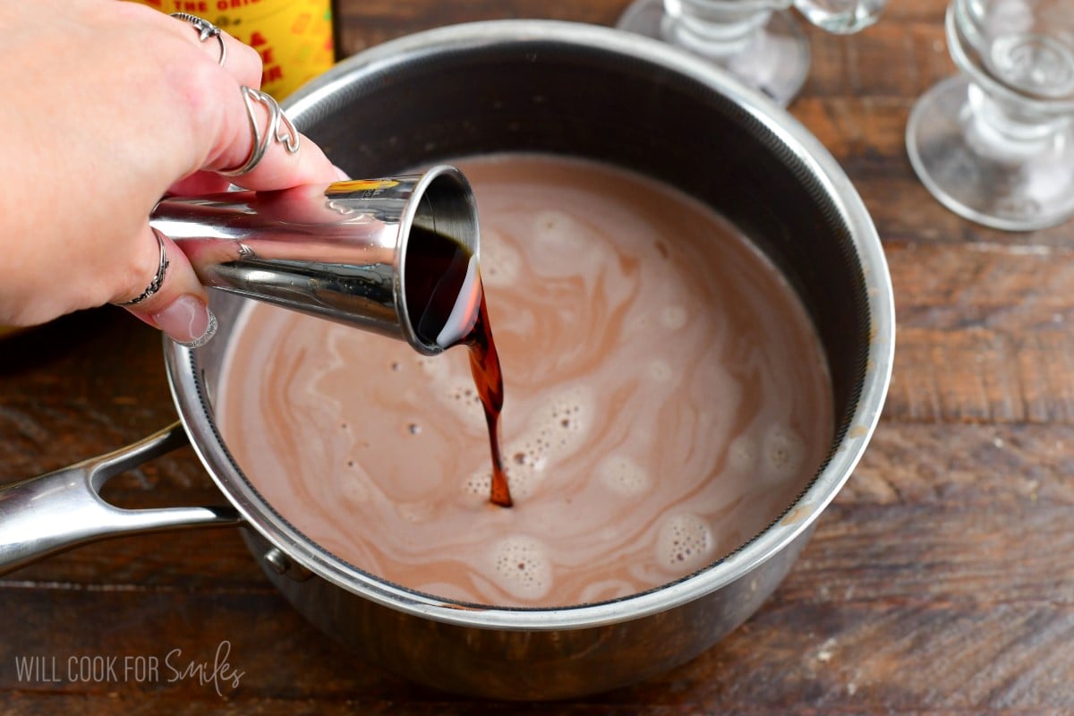 Adding kalua to the pot of hot chocolate that is sitting on a wood surface.