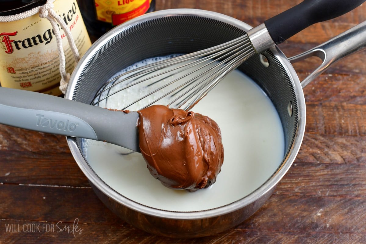 Adding nutella to pot of milk with a spoon and with a whisk sitting in the pot.