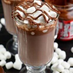 Nutella hot chocolate in a glass on a wood surface with mini marshmallows around the bottom of the glass.