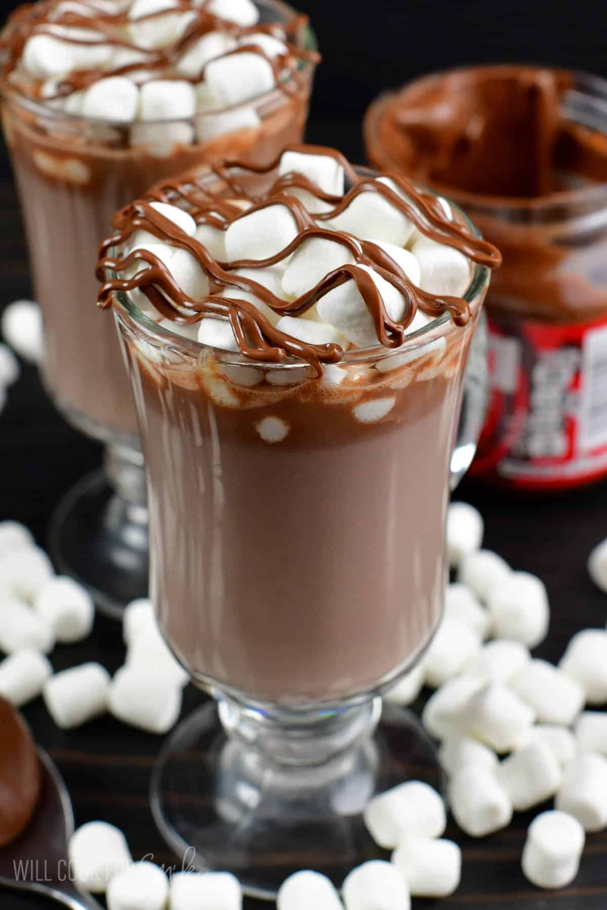 Nutella hot chocolate in a glass on a wood surface with mini marshmallows around the bottom of the glass.