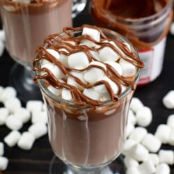 Nutella hot chocolate in a glass with marshmallows on a wood surface with marshmallows around the bottom.