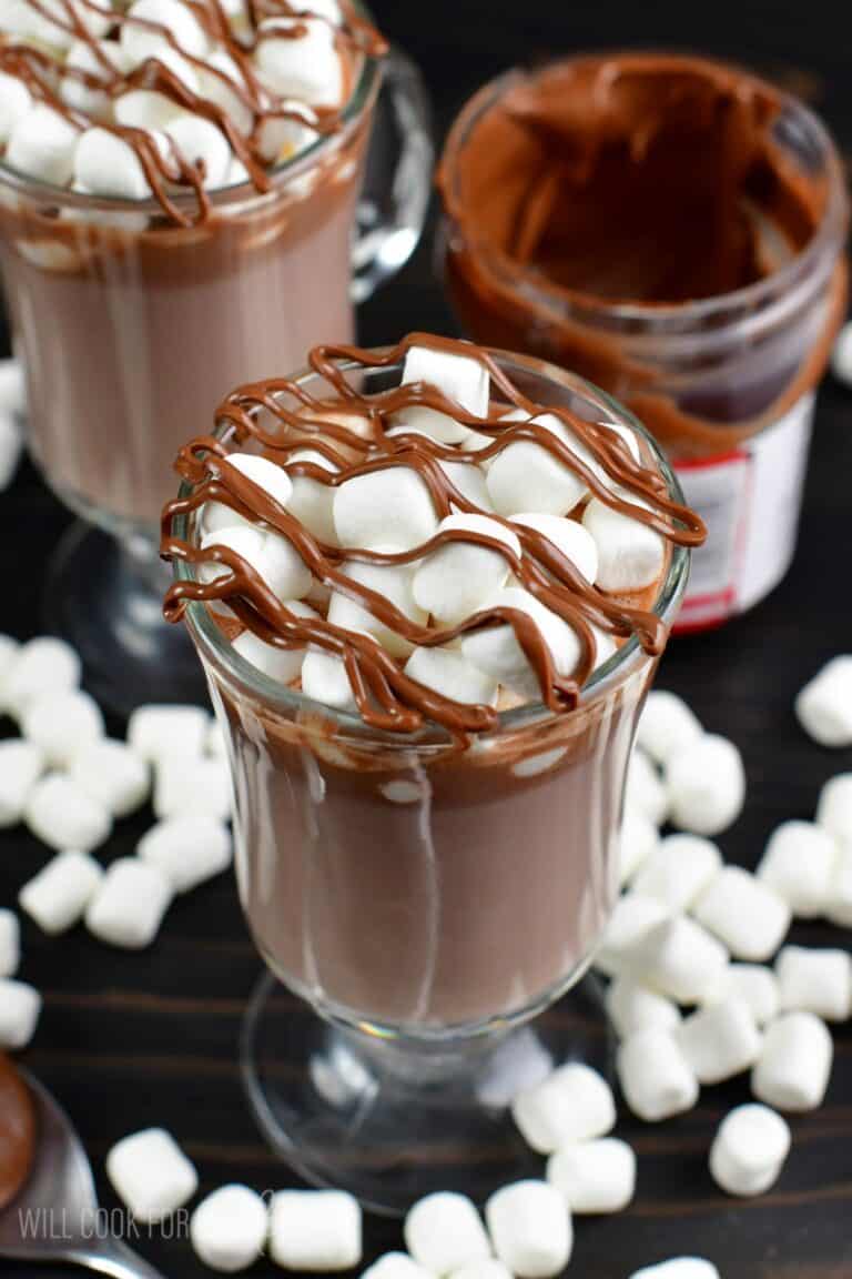 Nutella hot chocolate in a glass with marshmallows on a wood surface with marshmallows around the bottom.