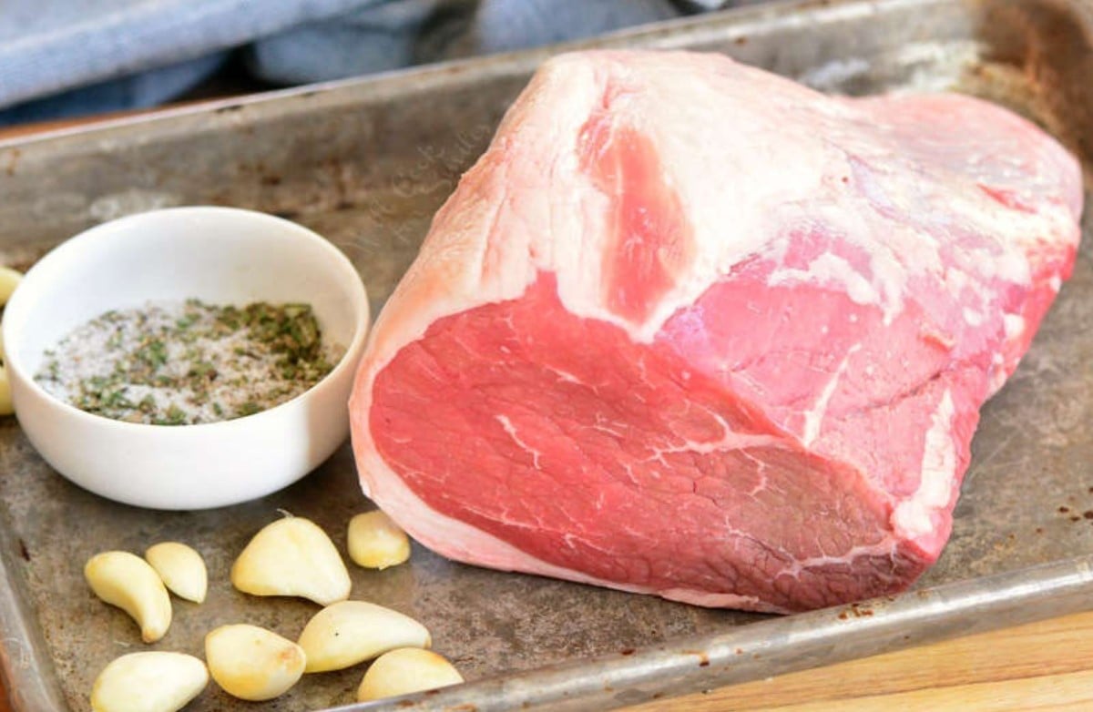 uncooked roast beef on a baking sheet with garlic and seasoning next to it.