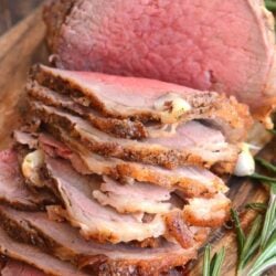 Roast beef sliced thinly on a cutting board with rosemary to the side of it.