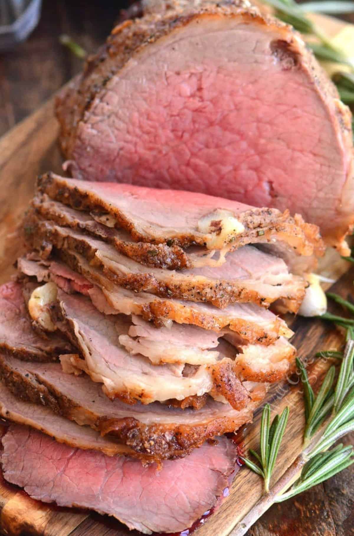 Roast beef sliced thinly on a cutting board with rosemary to the side of it.