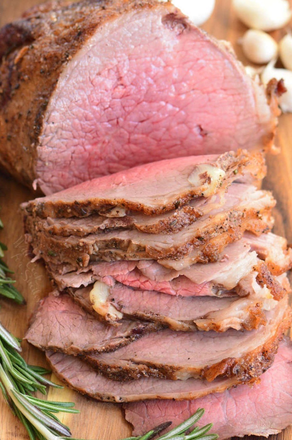 sliced roast beef on a wood cutting board with garlic and rosemary around it.