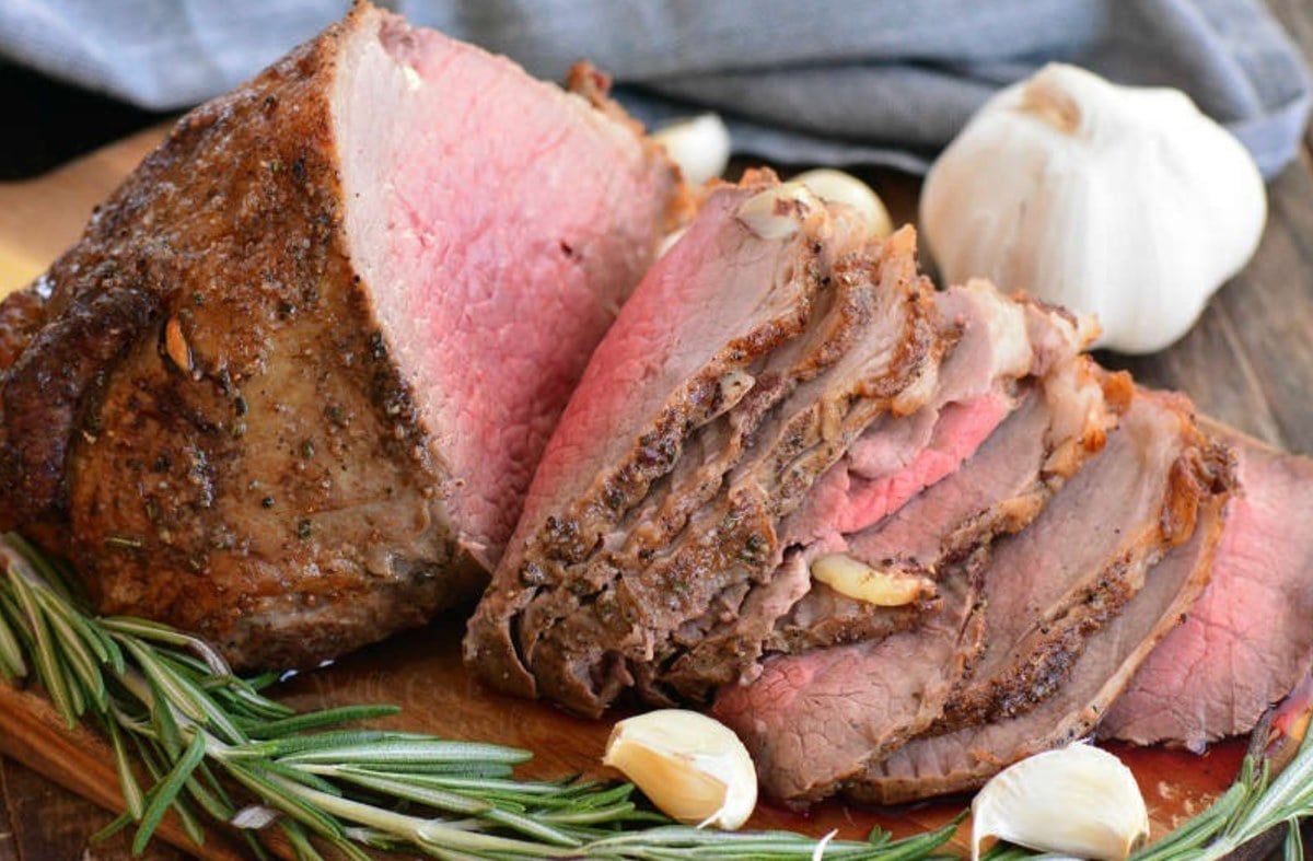 Cooked roast beef on a cutting board with slices in it and rosemary and garlic around it.