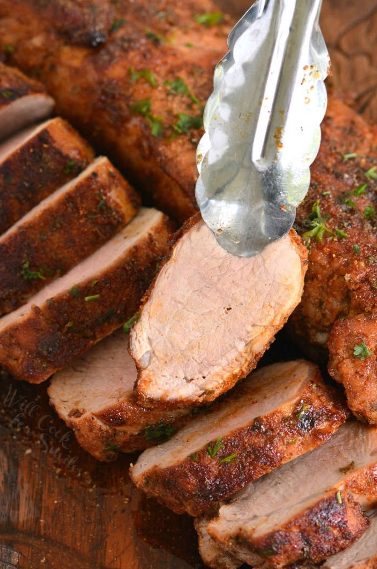 Two roasted pork tenderloins one sliced and one unsliced holding a slice with a pair of tongs.