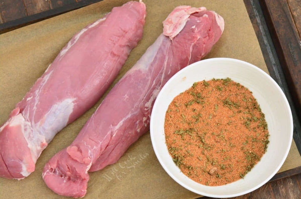 two pork tenderloins uncooked on a parchment lined baking sheet with a bowl of seasoning next to it.