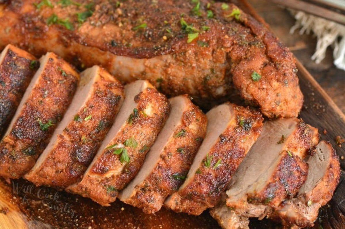 Two roasted pork tenderloins one sliced and one unsliced on a wood cutting board.