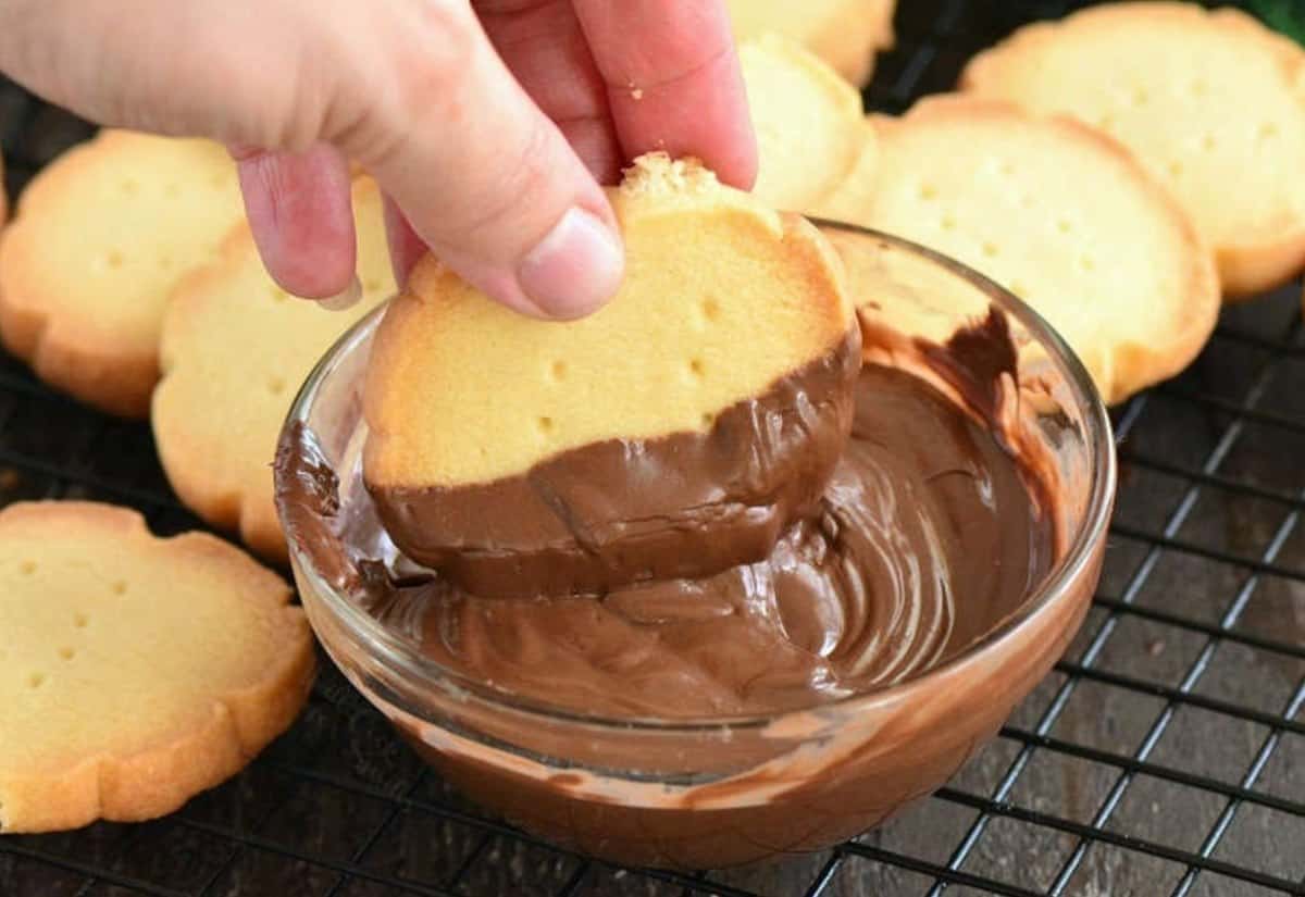 Dipping a shortbread cookie in melted chocolate that is in a glass bowl on a wire rack.