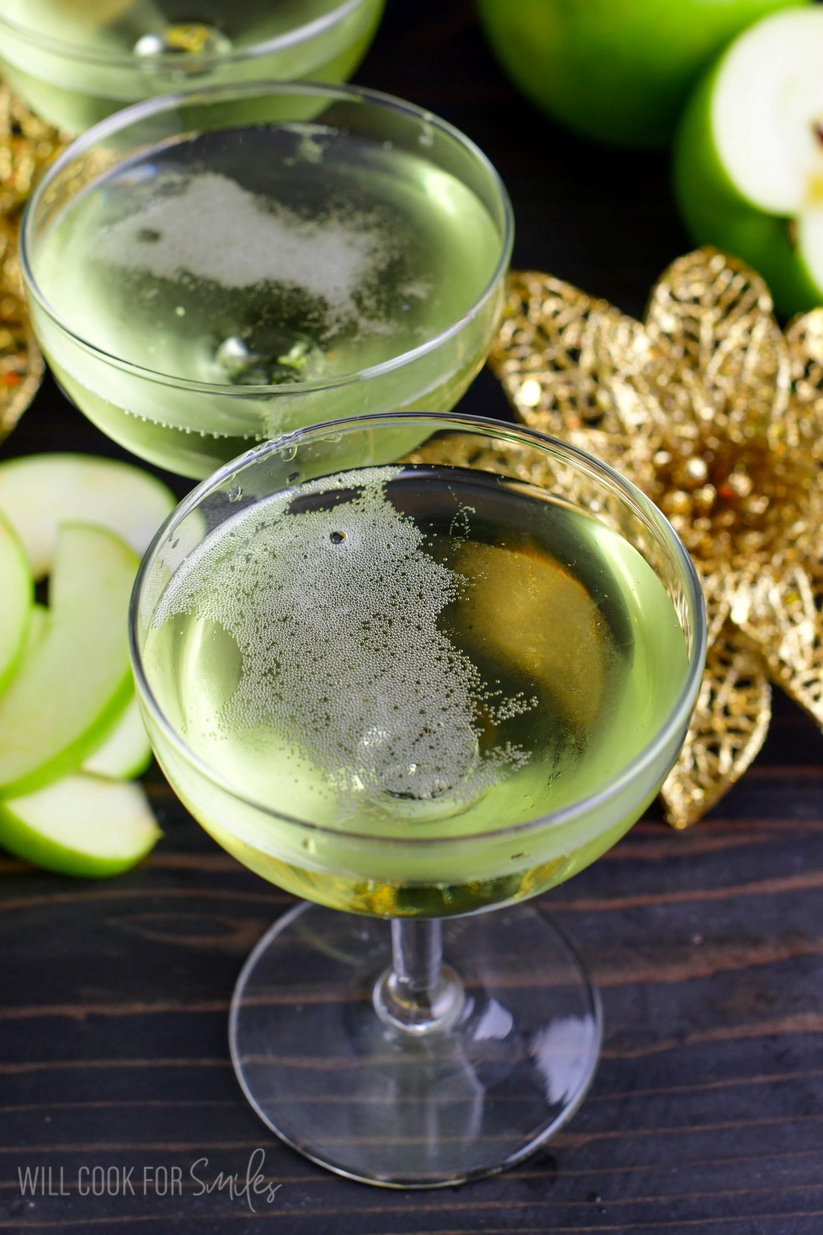 two glasses of sour apple champagne cocktail in a martini glass with gold flowers and apples.