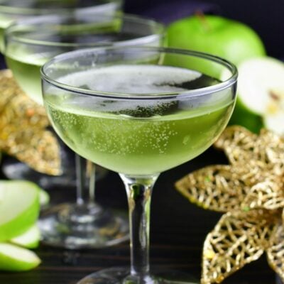 bubble sour apple champagne cocktail in a martini glass next to gold flowers and apples.