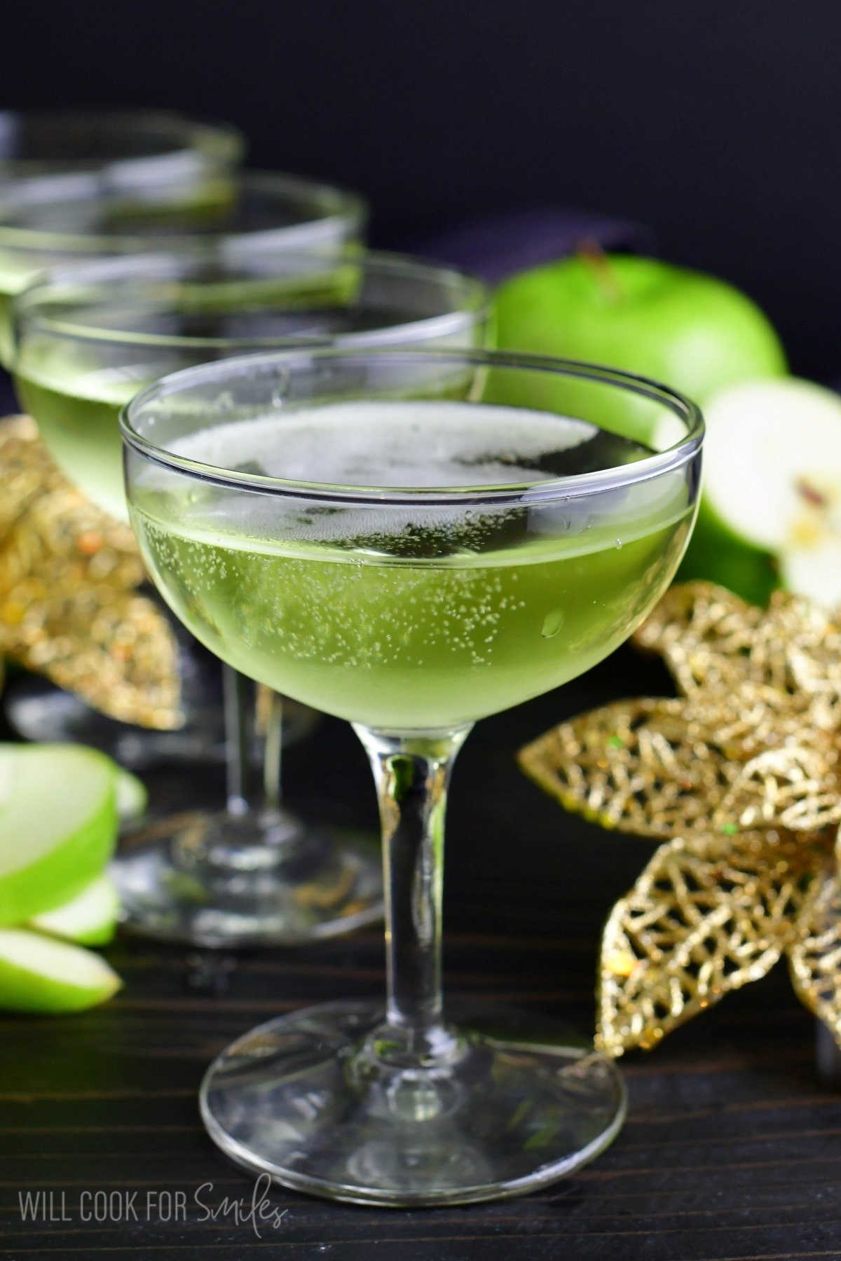 bubble sour apple champagne cocktail in a martini glass next to gold flowers and apples.