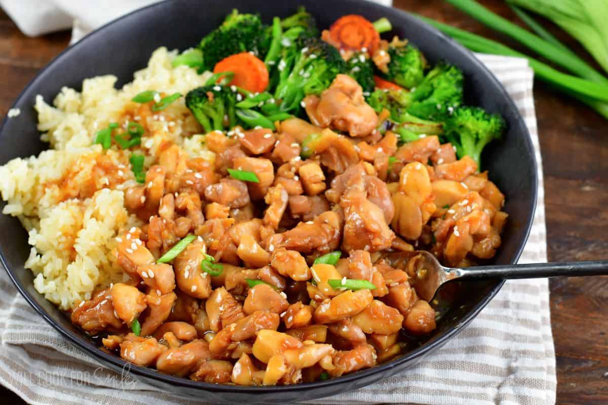 chicken teriyaki with rice, veggies, green onion and a fork in a black bowl.