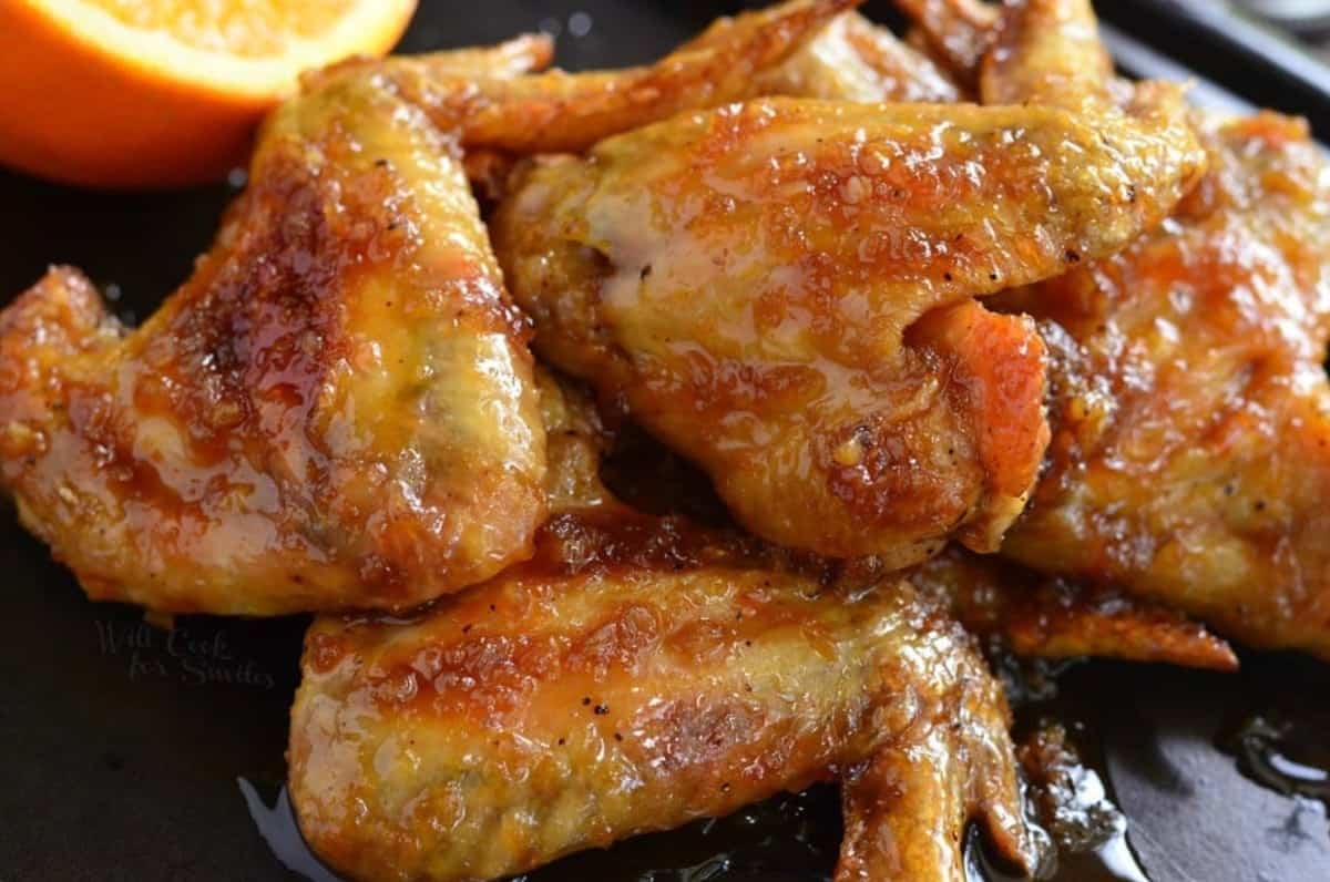 Chicken wings with orange glaze on them stacked up on a baking sheet.