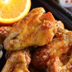 A stack of orange glazed chicken wings on a baking sheet with an orange on the side.